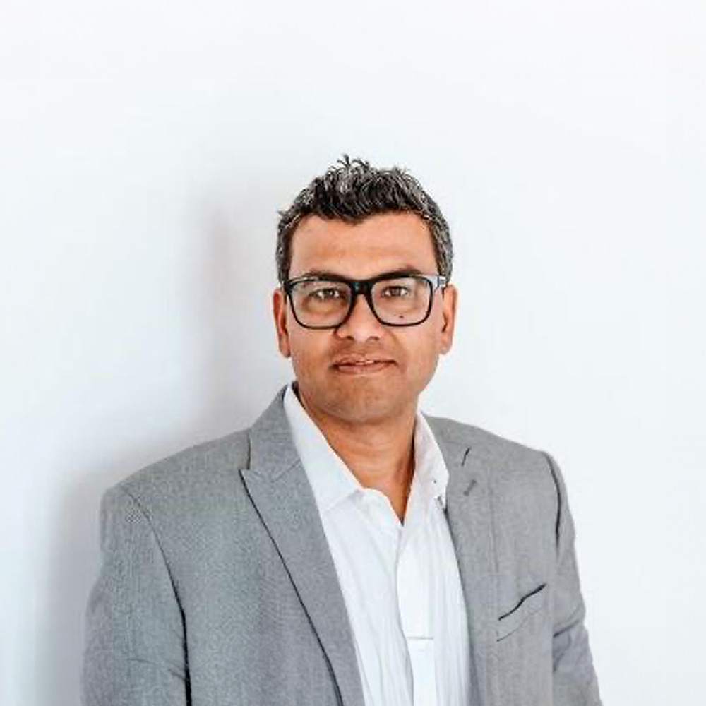 A man wearing glasses and a suit stands against a white wall, posing for a portrait with a neutral expression.