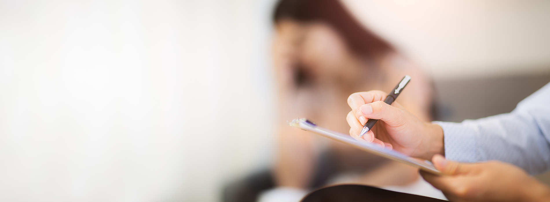 The image shows a person writing on a tablet with a stylus while seated at a table, with another tablet and pen in front of them, suggesting an environment focused on note-taking or reviewing documents.