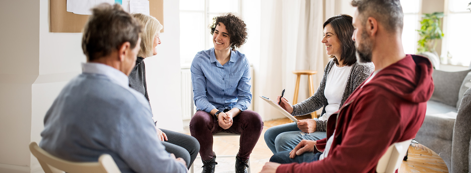 The image shows a group of people sitting around a table, engaged in conversation, with some standing and others seated, all appearing to be in a casual meeting or discussion.