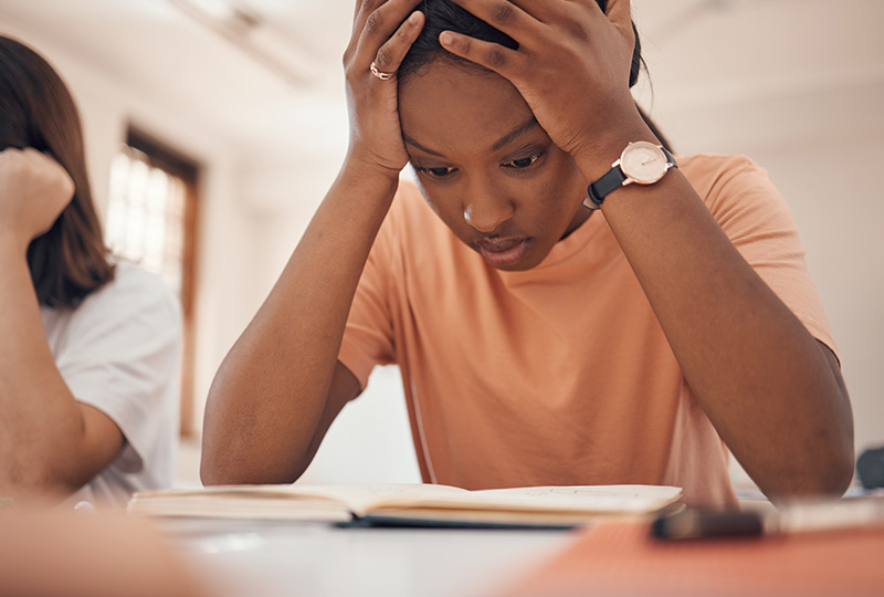 The image shows a person with their head resting on their hand, appearing distressed or overwhelmed, sitting at a desk with an open book, while another person looks on with a concerned expression.