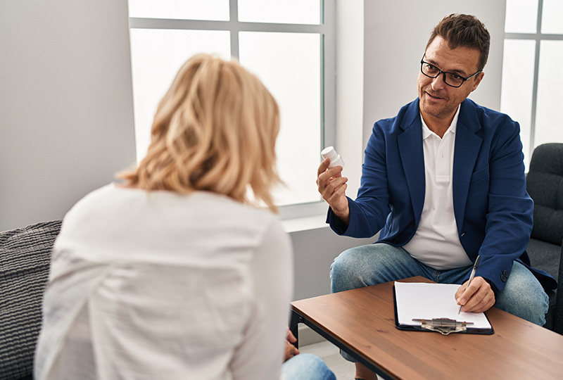 A man in a blue suit sitting at a table with a woman who is holding a tablet. The man appears to be listening attentively while the woman is presenting something on the tablet.