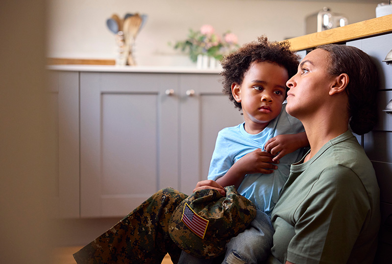 The image depicts a woman in military uniform holding a young child, both seated indoors with a warm, intimate atmosphere.