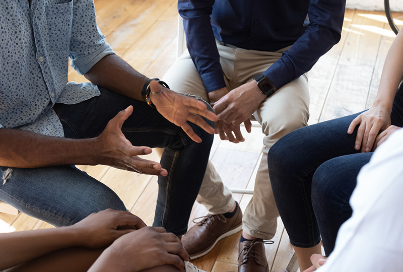 The image depicts a group of four people sitting around a table with their hands clasped together, engaged in conversation. They are dressed casually, suggesting an informal meeting or discussion.