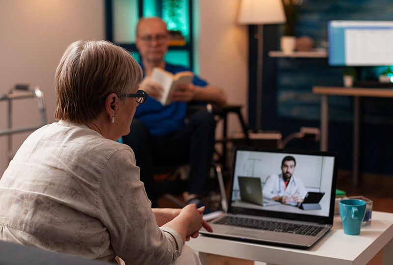 The image shows two individuals sitting at a table with a laptop, engaged in a video call with another person on the screen. They are indoors, possibly in a living room setting, and there s a book visible nearby.