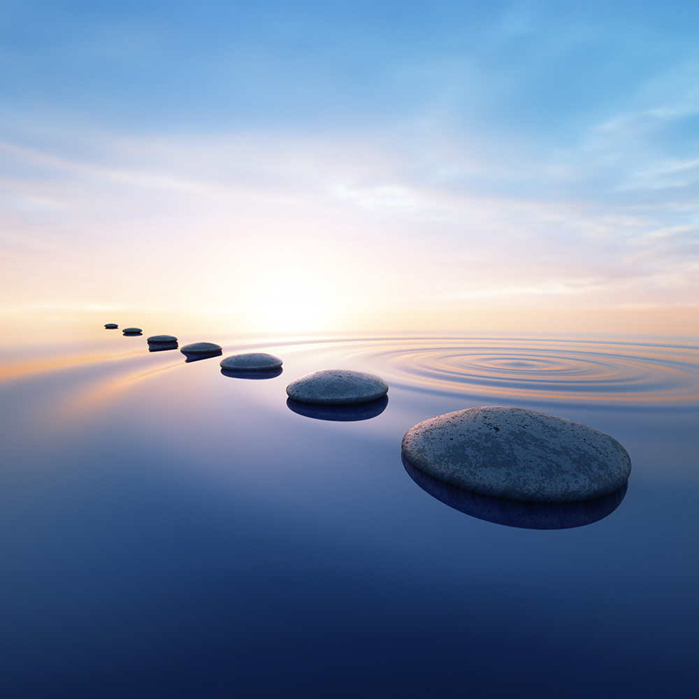 The image shows a serene beach scene at sunset with a long exposure photograph capturing the movement of water creating concentric circles around a set of three stones on a pathway leading into the distance, evoking a sense of tranquility and contemplation.