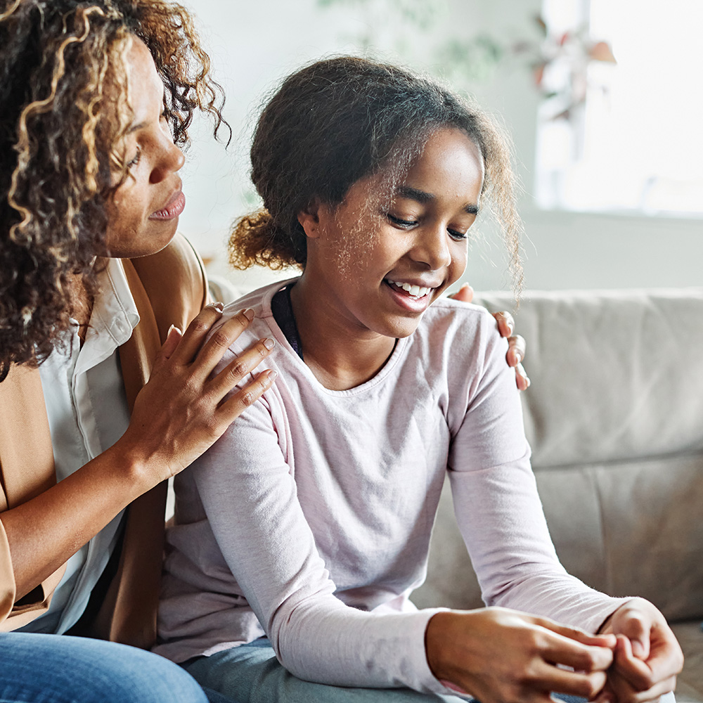 The image shows a woman sitting on a couch with her hands on a young girl s shoulders, both smiling at the camera.