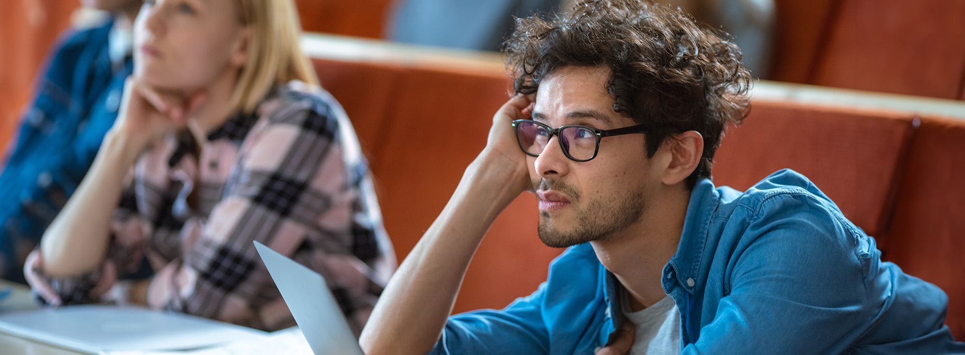 A man with glasses, wearing a blue shirt, sits at a desk in a classroom setting, looking down with his chin resting on his hand.