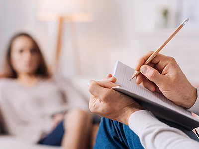 A person sitting on a couch with their feet up, holding a notebook and pen, writing while looking at another person who is seated across from them.