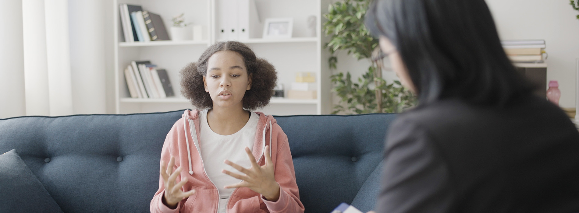 The image shows two individuals engaged in a conversation, with one person seated on a couch and another standing, both appearing to be in an indoor setting.