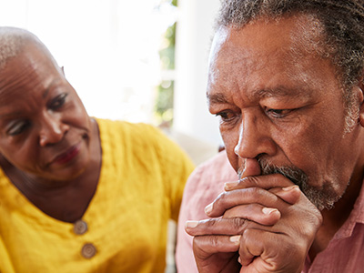 The image shows two older adults, possibly family members, with one person looking down at their hands clasped together, seemingly concerned or contemplative, while another person looks on with a focused gaze.