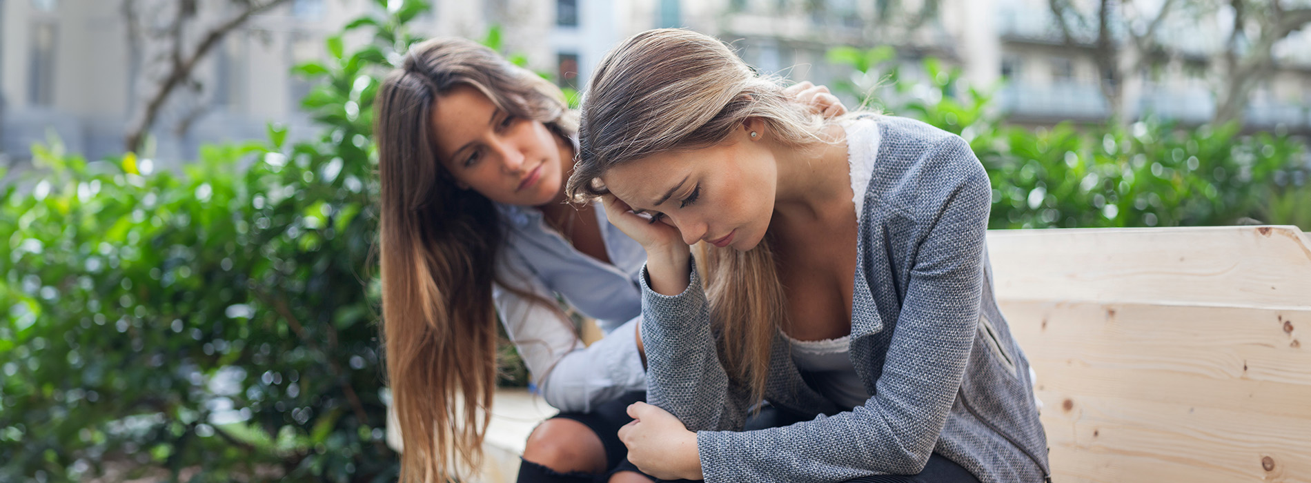 A woman sitting on a bench with her head resting on her hand, while another woman stands beside her, both looking downward, seemingly engaged in conversation.