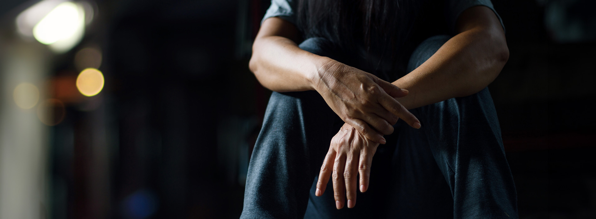 The image shows a person sitting with their hands clasped together, facing downwards, against a dark background with a blurred street scene visible in the background.