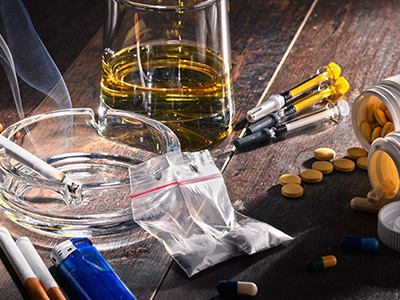 The image shows a table with various items associated with substance abuse, including an open bottle of alcohol, cigarettes, pills, and paraphernalia, suggesting a scene of drug use.